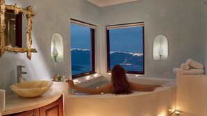 A woman relaxing in a bathtub with a view of a mountainous landscape through two windows. - Maison Des Lys (Santorini)