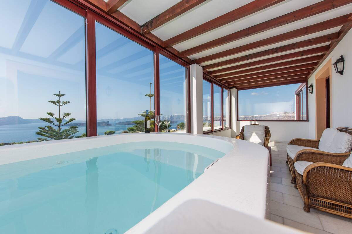 A modern infinity pool with a clear view of the sea and mountains, wooden ceiling beams, wicker furniture, and a glass-enclosed patio area.