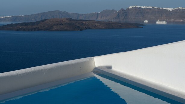 A clear view of a swimming pool with a white edge, a calm blue sea, and a distant island with mountains. - Maison Des Lys (Santorini)