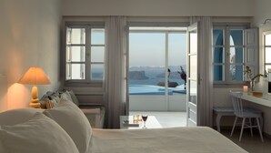 A hotel room with a bed, a desk, a chair, a lamp, and a view of the sea. - Maison Des Lys (Santorini)