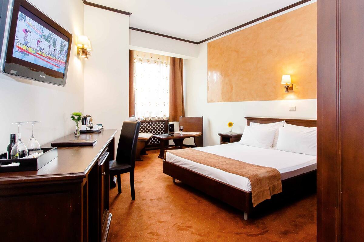 Una habitación de hotel con cama, escritorio, silla, televisión y ventana con cortinas.