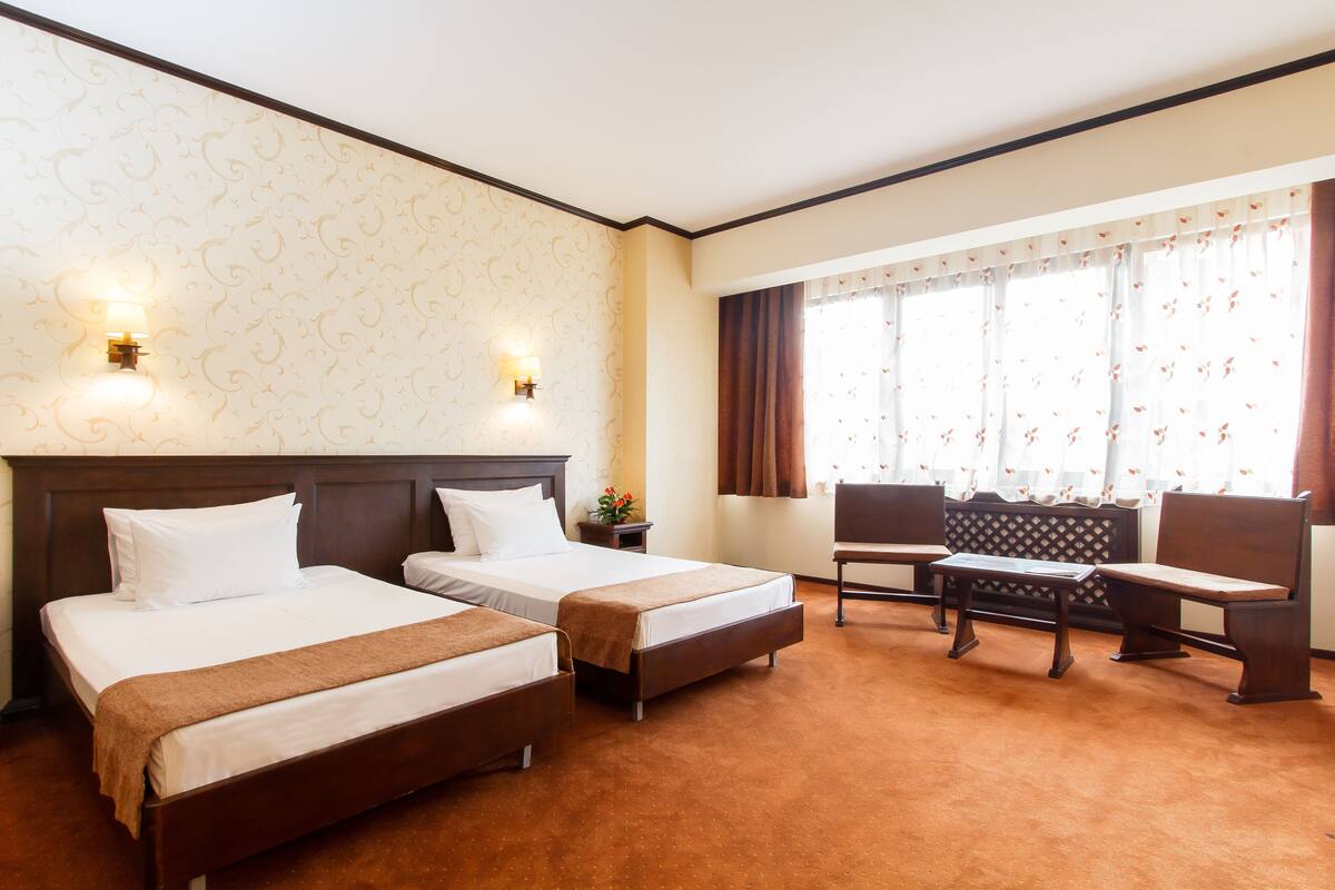 Habitación de hotel con dos camas, un banco, una mesa y un ventanal con cortinas.