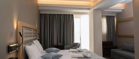 A hotel room with a large bed, a sofa, and a desk. There is a window with curtains, a wall-mounted TV, and a bedside table with a lamp and a coffee maker.