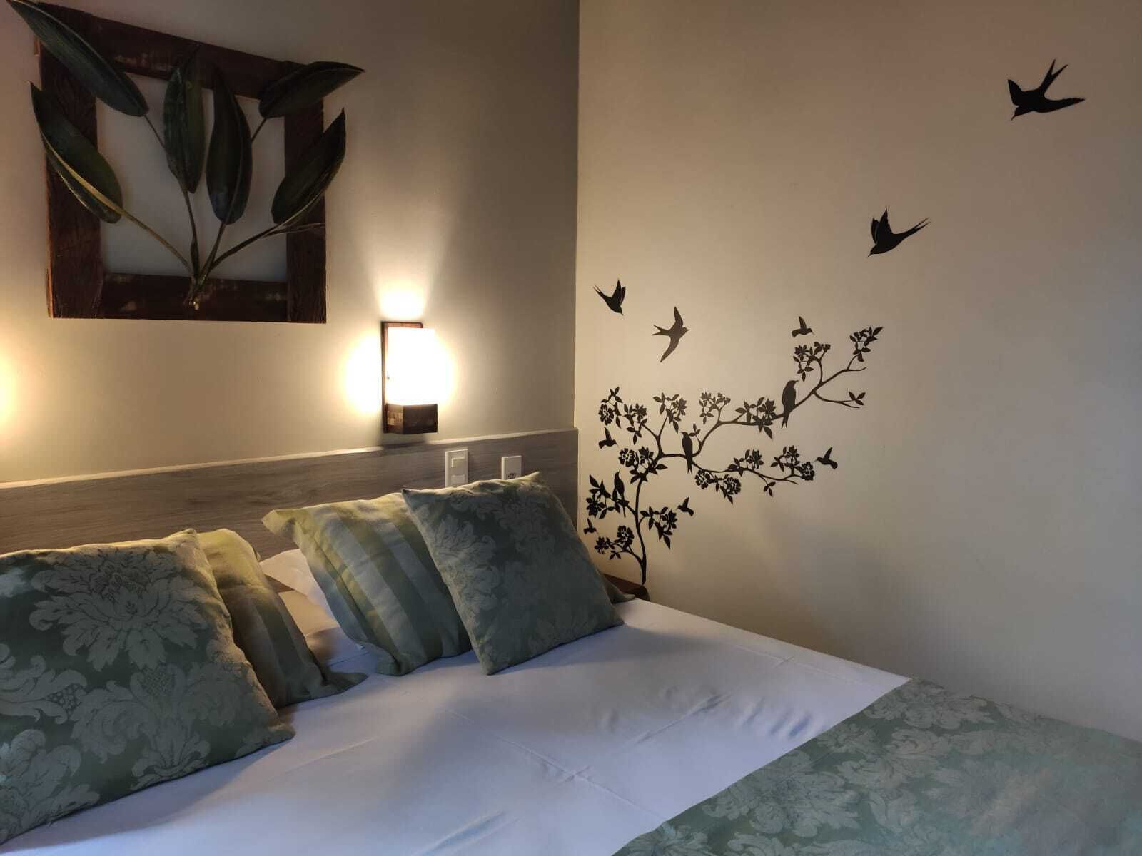 A hotel room with a bed, pillows, a wall-mounted lamp, and a wall decal of birds and flowers.