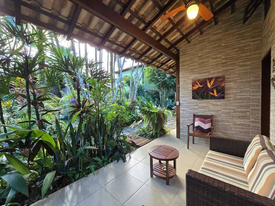 A covered patio with a wicker sofa, a small round table, and a chair. The patio is surrounded by lush tropical plants and a brick wall.
