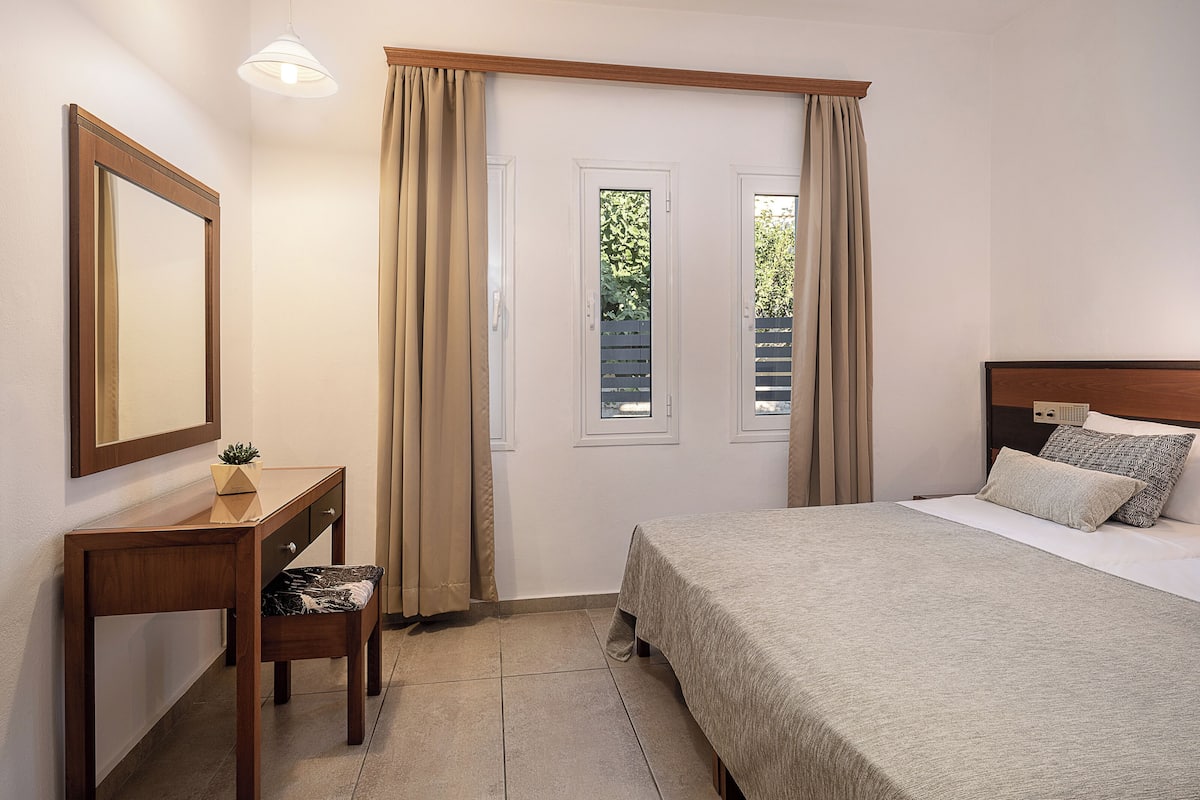 A bedroom with a bed, a desk with a chair, a mirror, and two windows with curtains.