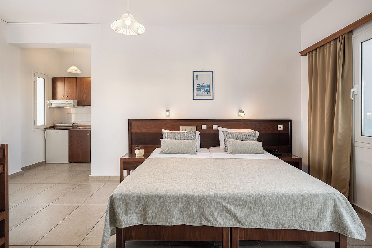 A hotel room with a bed, bedside tables, a nightstand, a small kitchen area, and a window with curtains.