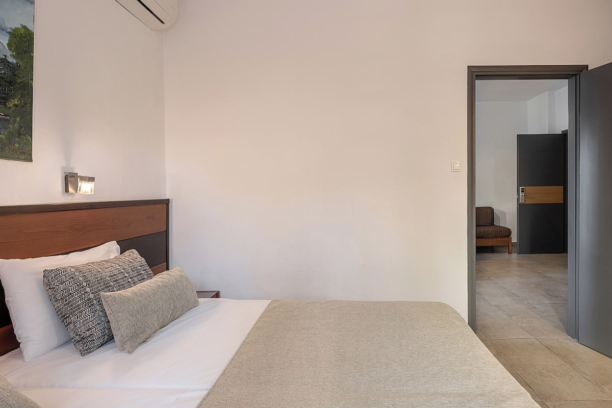 A bedroom with a bed, a nightstand, a wall-mounted air conditioner, a painting, and a door leading to another room.