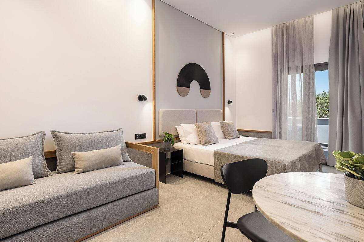 A modern hotel room with a sofa, a bed, a dining table, and a view of the outdoors.