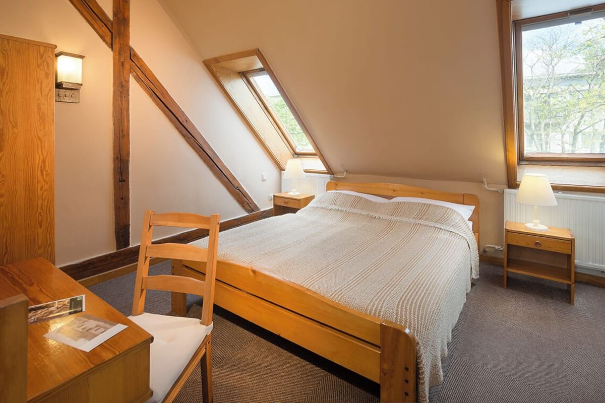 A bedroom with a wooden bed, a chair, a nightstand, a lamp, and a window with a view of trees.