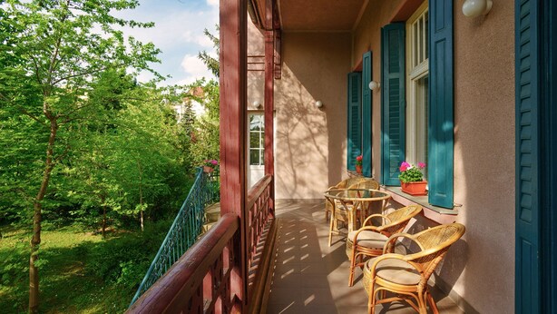 Un balcón con sillas de mimbre y una mesa, con vistas a un jardín exuberante y árboles.