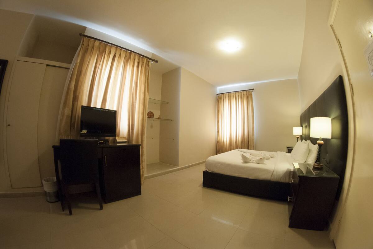 A hotel room with a bed, a desk with a television, a nightstand with lamps, and curtains.