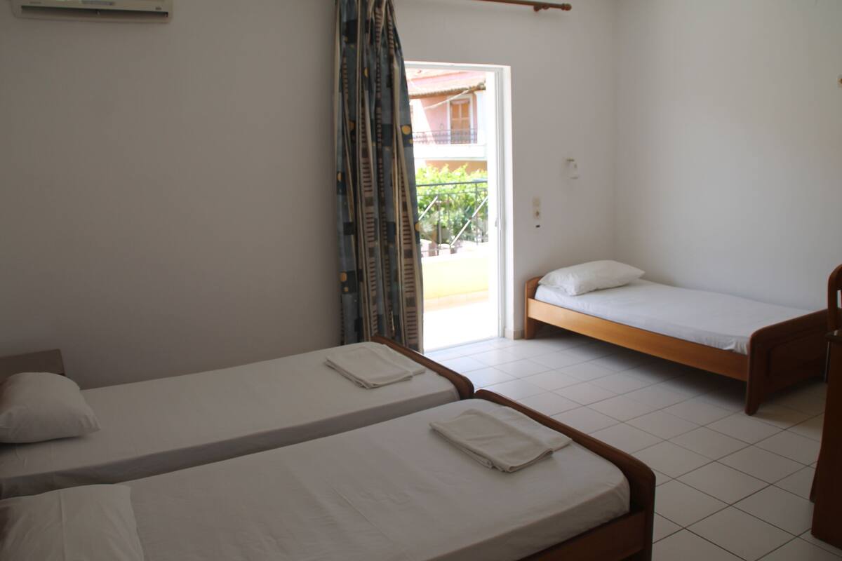A room with two single beds, a wooden headboard, a white ceiling fan, and a view of a building outside.
