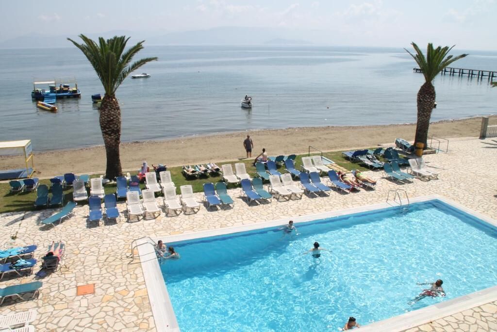 Zona de piscina con sillones de descanso y palmeras, con vista a una playa con barcos.