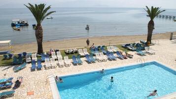 Zona de piscina con sillones de descanso y palmeras, con vista a una playa con barcos.