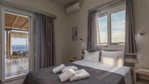Una habitación de hotel con cama, mesita de noche, ventana con cortinas, vista al mar y balcón con sillas.