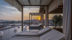 Una terraza en la azotea con vistas al atardecer sobre un paisaje urbano, que incluye sillones de descanso y una cubierta de madera.
