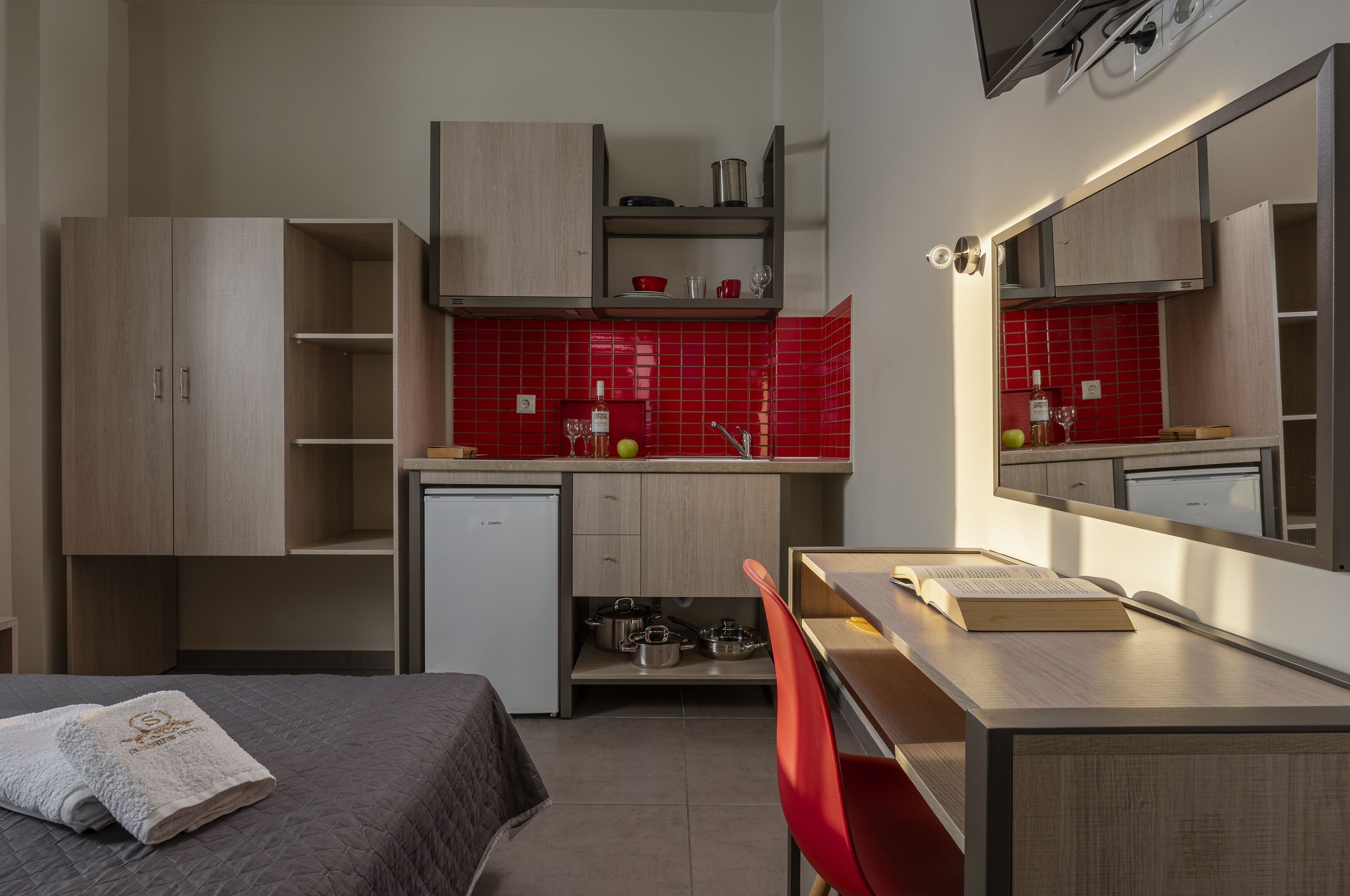 Cocina compacta con microondas, pileta y muebles de madera. Incluye un refrigerador pequeño, una silla roja y una cama con una toalla.