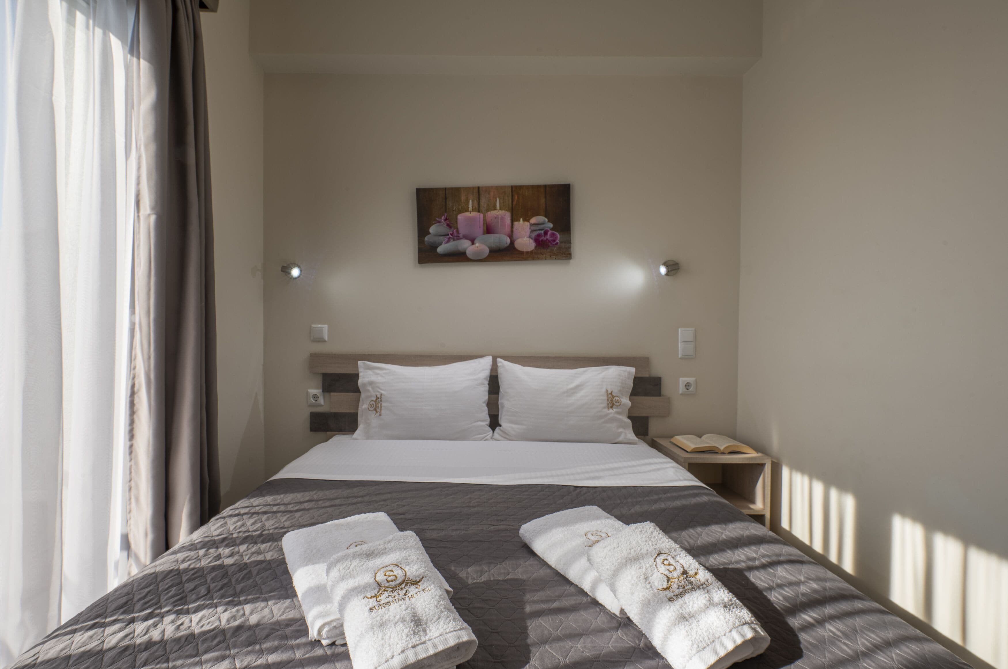 A hotel room with a large bed, two pillows, a nightstand with a book, and a painting on the wall.