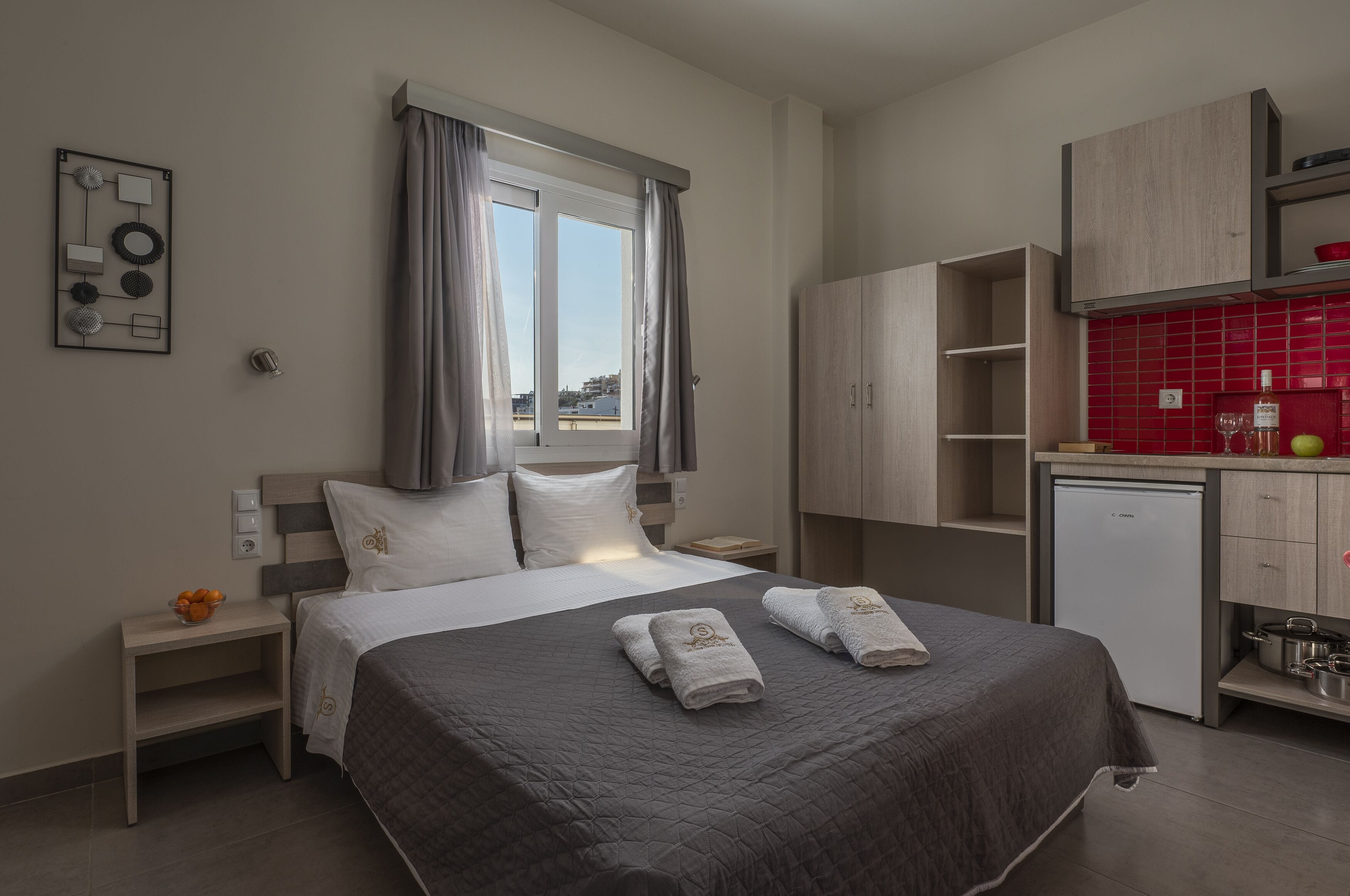 A modern hotel room with a large bed, a small kitchenette, and a view through the window.