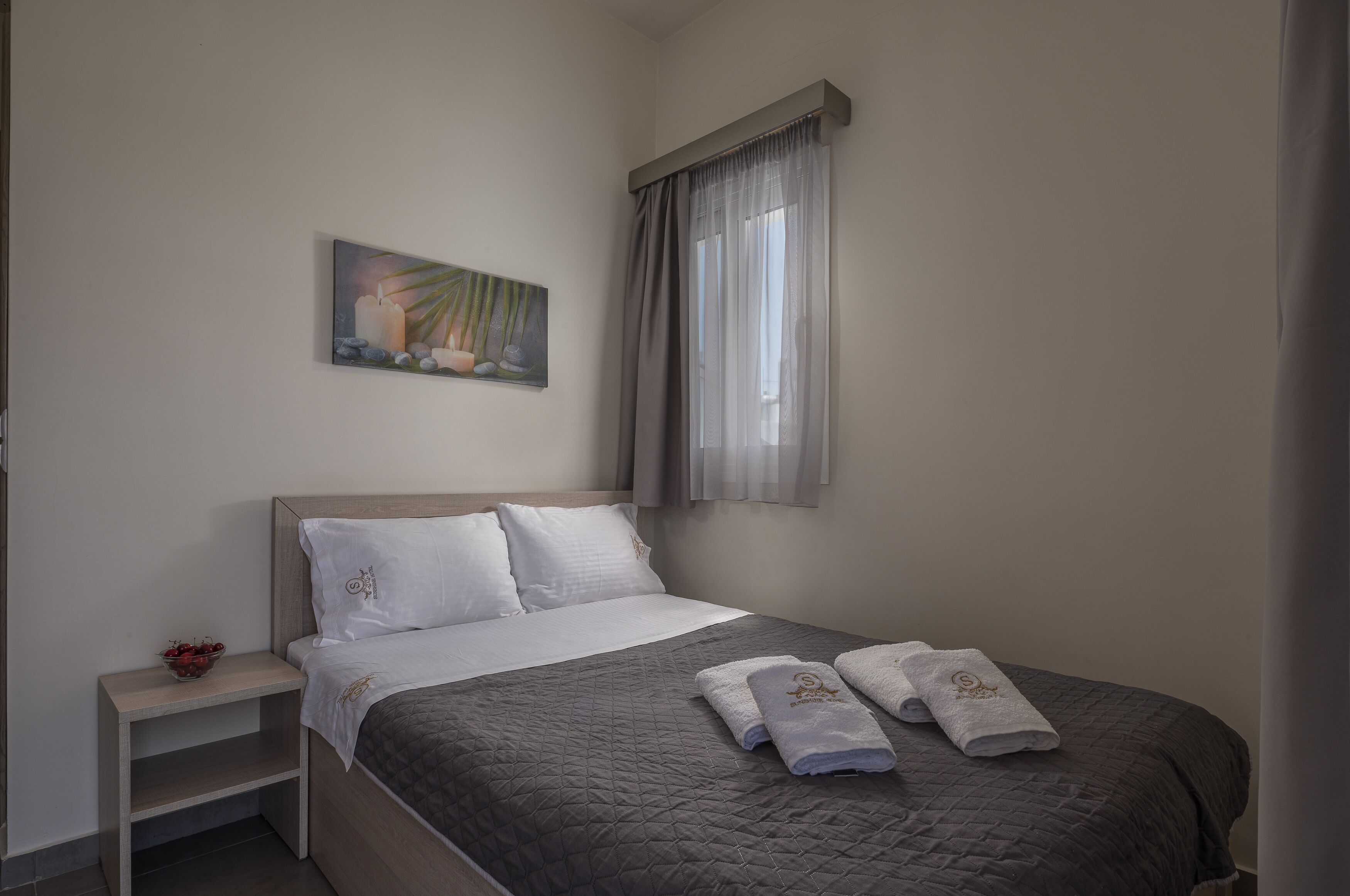 A neatly made bed with a gray quilt, two white pillows, and two towels with a logo. There's a small bedside table with a bowl of fruit and a painting of a candle and flowers on the wall.