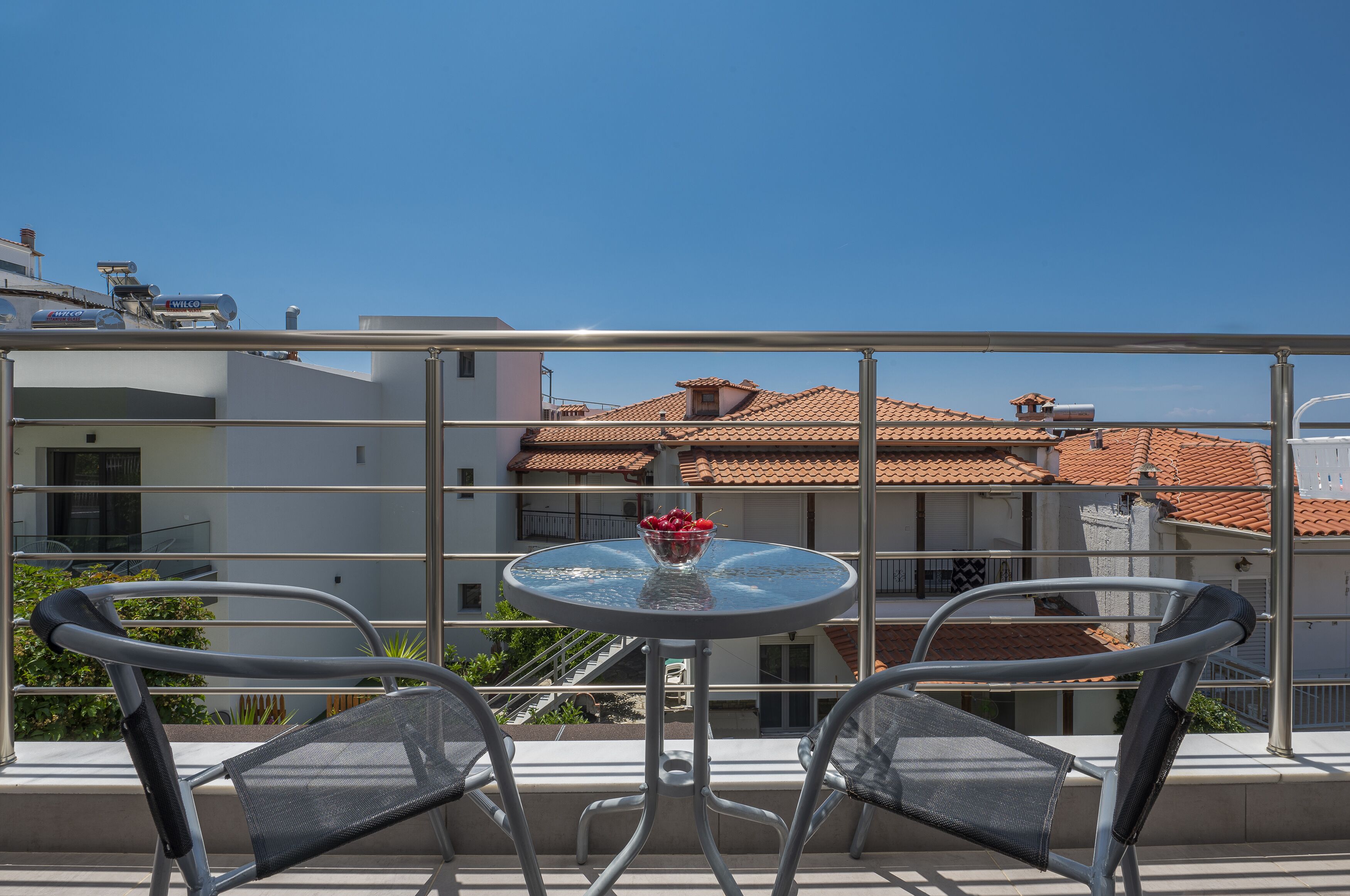A balcony with a round glass table and two chairs, overlooking a building with a tiled roof.