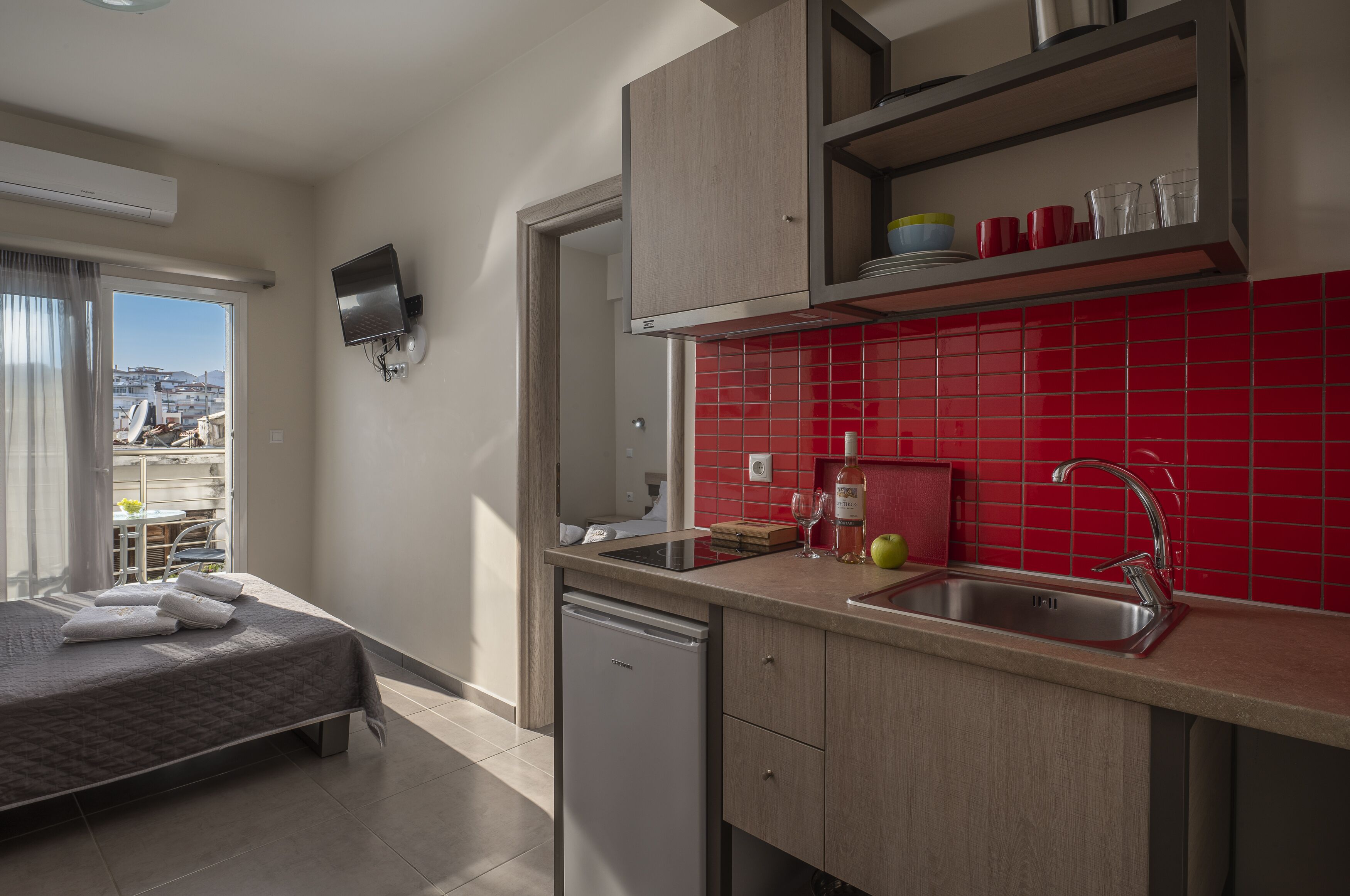 A modern hotel room with a bed, a kitchen area with red tiles, a dishwasher, and a balcony with a view.