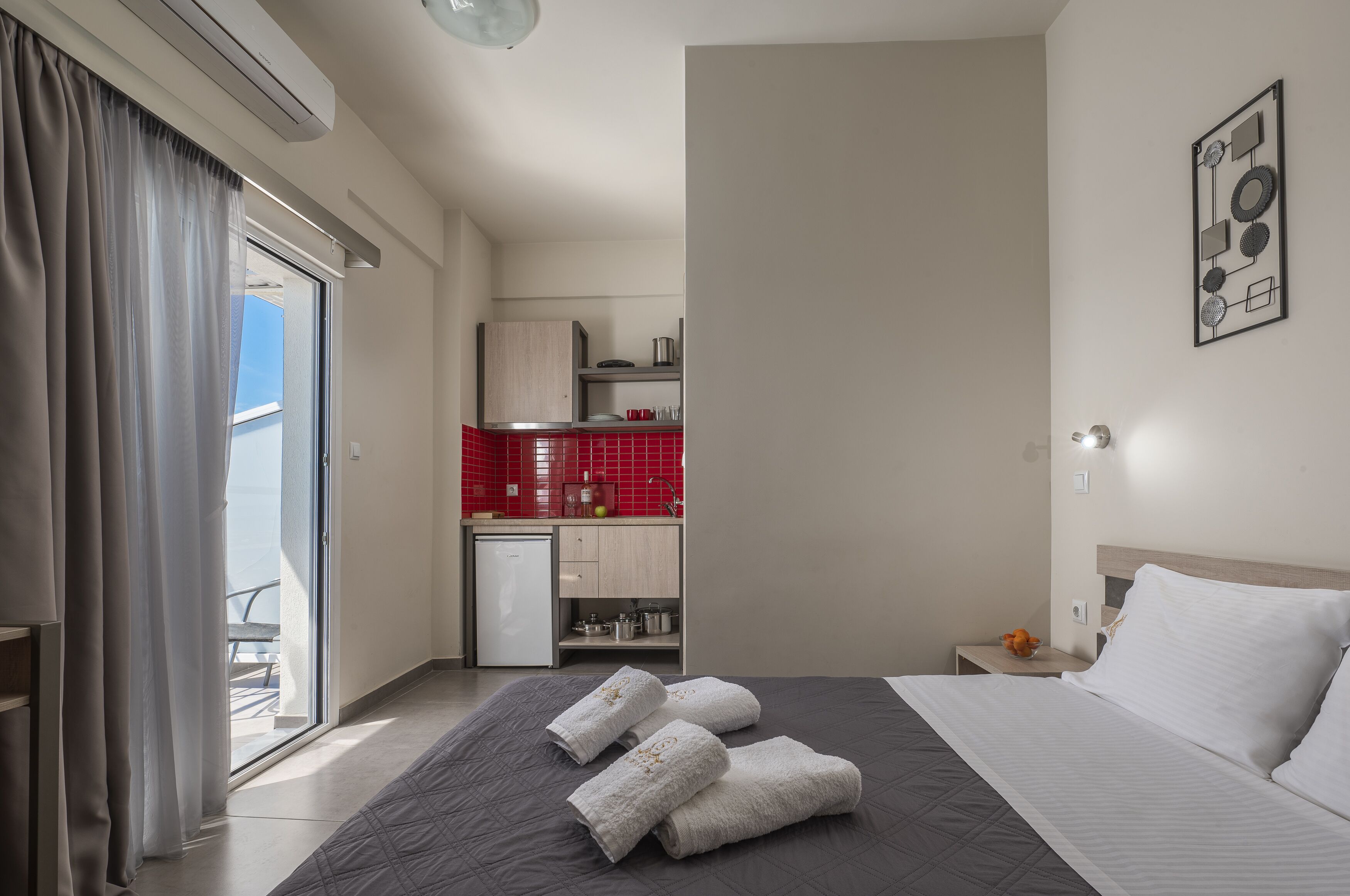 Une chambre d’hôtel avec un lit, une table de chevet, une petite table avec un vase, une vue sur le balcon et une kitchenette carrelée de carreaux rouges.