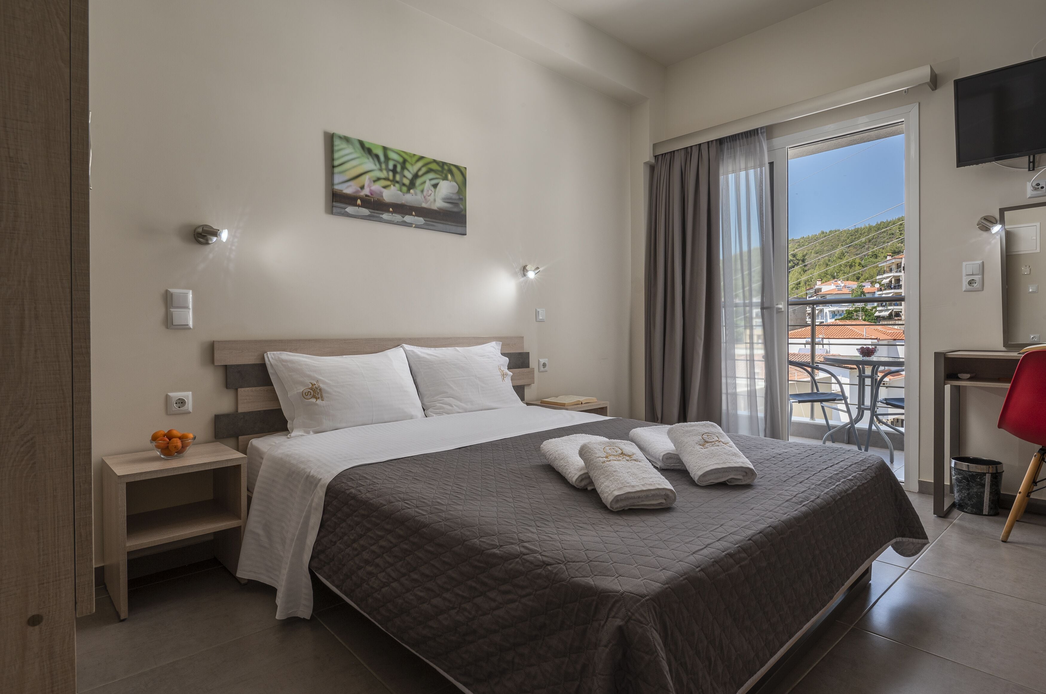 Ein Hotelzimmer mit einem großen Bett, einem Schreibtisch mit Stuhl, einem Fernseher und einem Balkon mit Blick auf die Stadt.