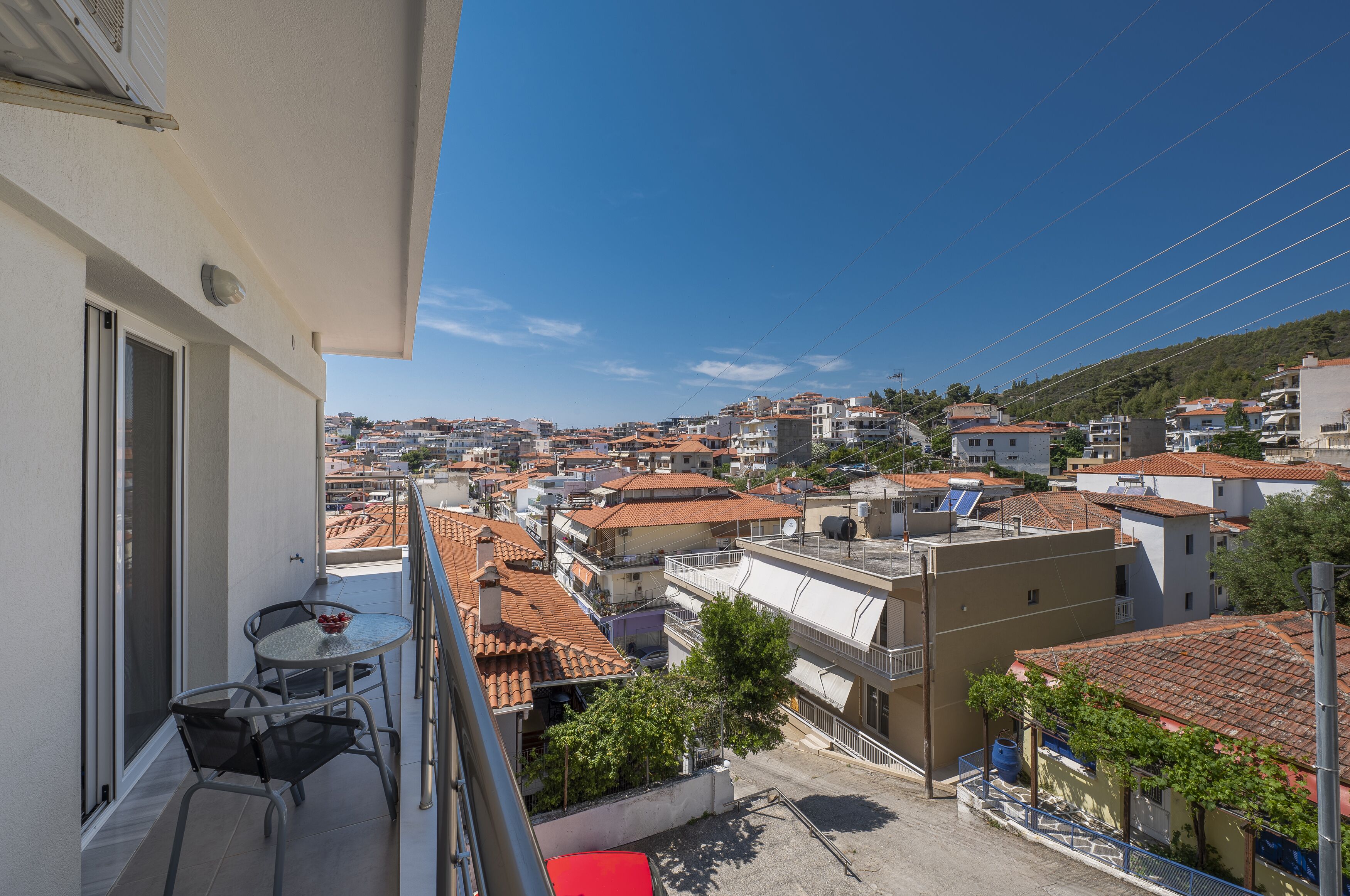 A balcony with a table and chairs, overlooking a residential area with red-tiled roofs and a clear blue sky.