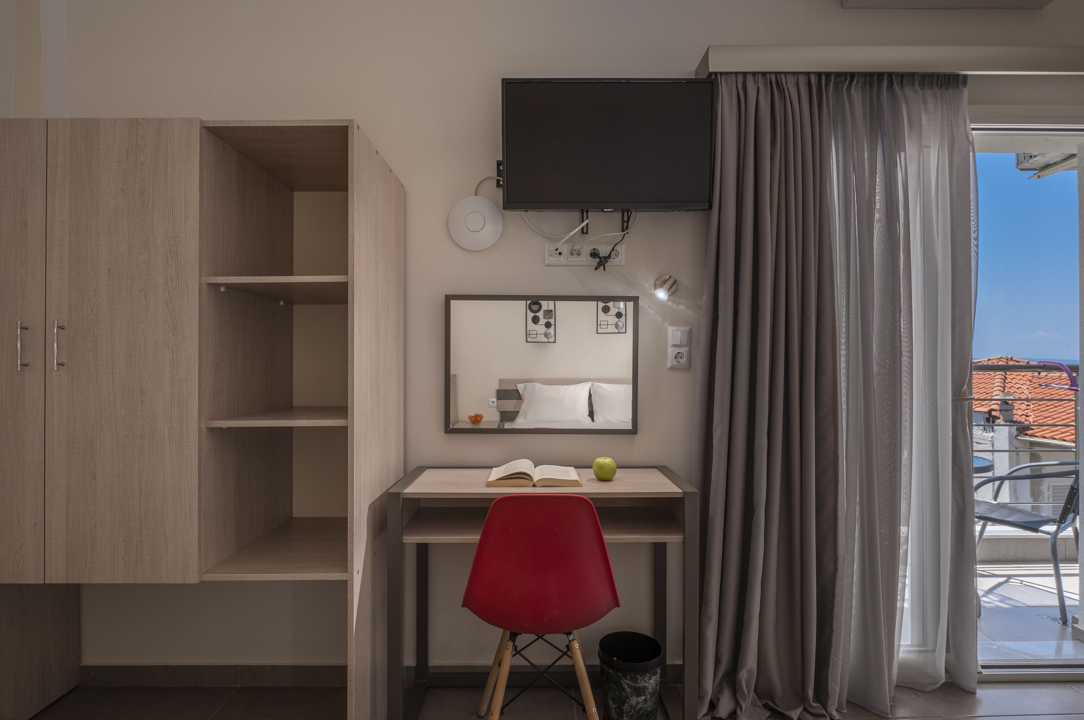 A hotel room with a wooden wardrobe, a desk with a red chair, a flat-screen TV, a mirror, and a window with curtains.