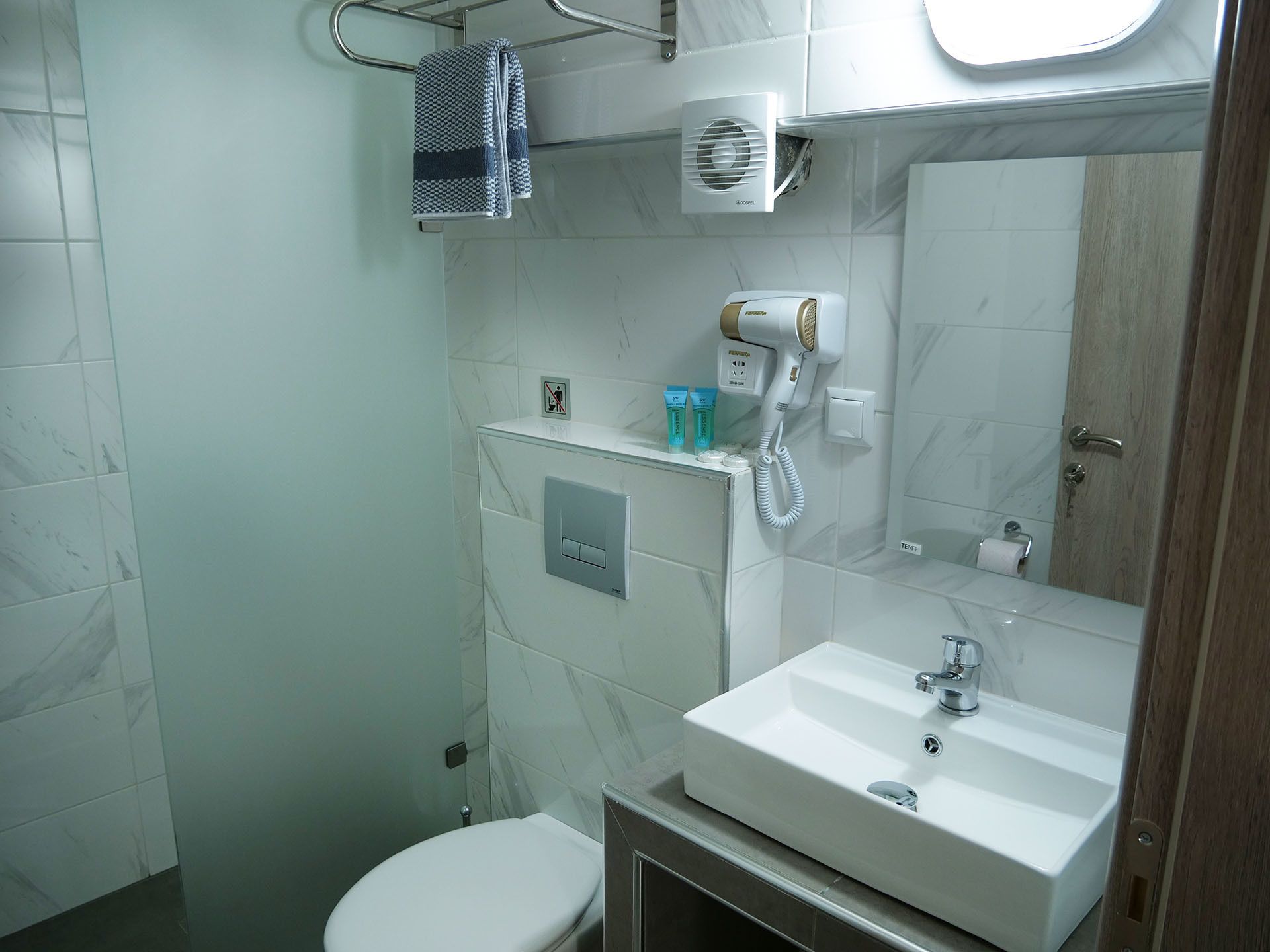 Une salle de bain avec des toilettes blanches, un lavabo avec un miroir, un porte-serviettes et un sèche-cheveux fixé au mur.