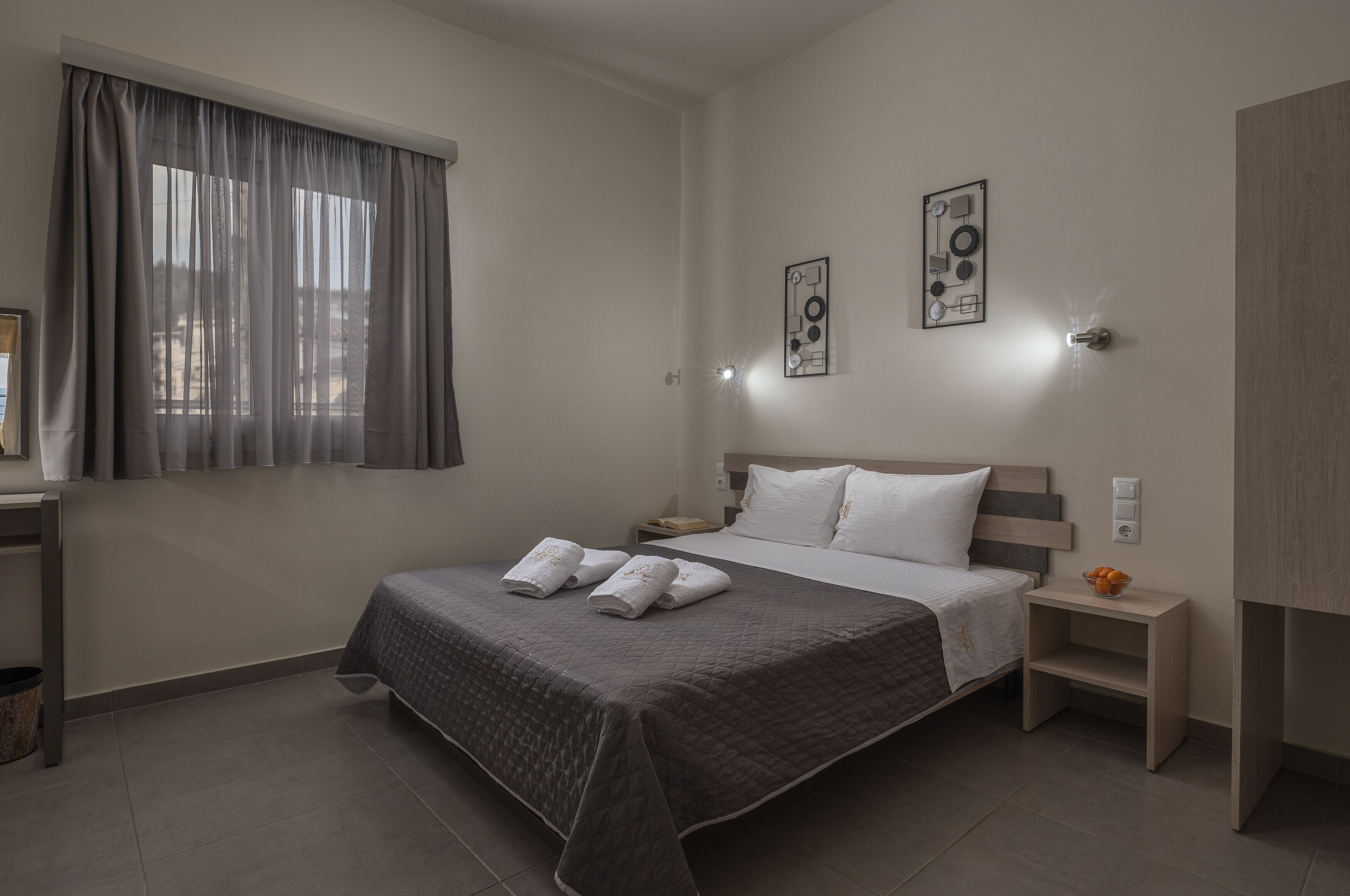 A neatly arranged bedroom with a bed, bedside tables, a nightstand, a wall-mounted light, and a window with curtains.