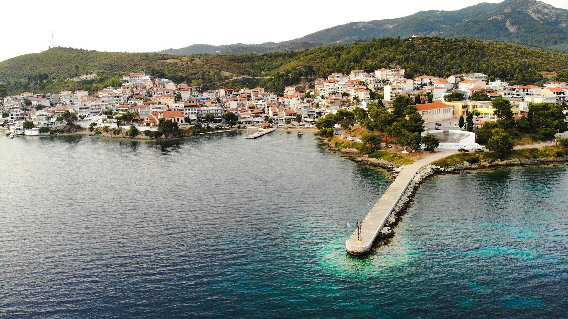 Un pueblo costero con un muelle que se adentra en el mar, rodeado de viviendas y zonas verdes.