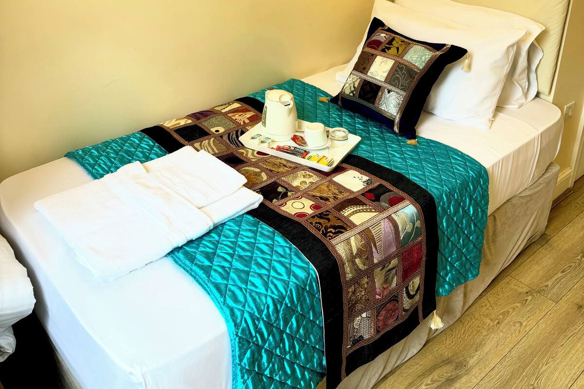 A single bed with a quilt, a bedside table with a cup and a tray, and a wall-mounted light.