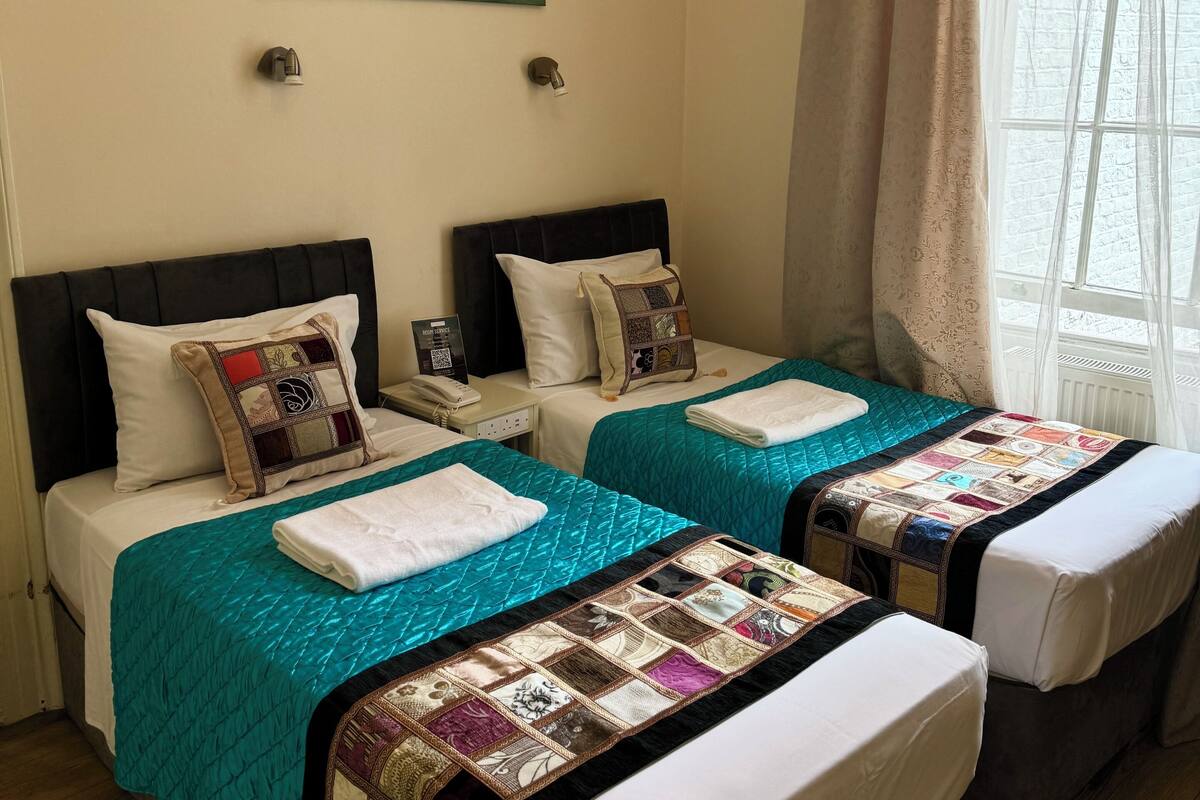 A hotel room with two beds, each with a teal quilt and a white pillow. There is a painting of a seascape on the wall, and a window with curtains.