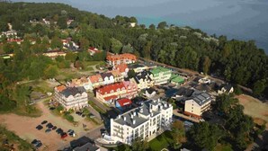 Vue aérienne d’une ville côtière avec différents bâtiments, dont un grand bâtiment blanc doté d’un toit vert, des maisons plus petites et un espace de stationnement.