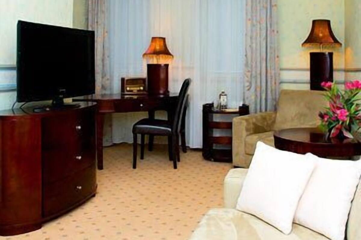 Een hotelkamer met een televisie, twee lampen, een bureau, een stoel, een bank en een decoratieve plant.
