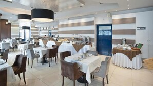 A modern dining area with multiple tables and chairs, a buffet setup, and a blue door in the background. - Trokadero Hotel (Delphi)