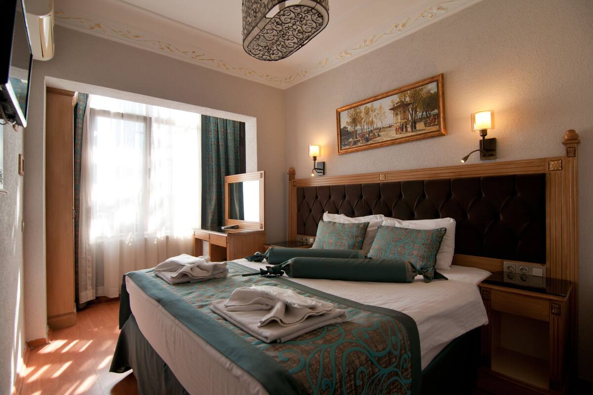 A hotel room with a large bed, a bedside table, a nightstand, a painting on the wall, and a window with curtains.