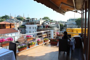 Une terrasse sur le toit, aménagée avec des espaces de détente en plein air, des plantes en pot et offrant une vue sur le paysage urbain, notamment sur une mosquée.