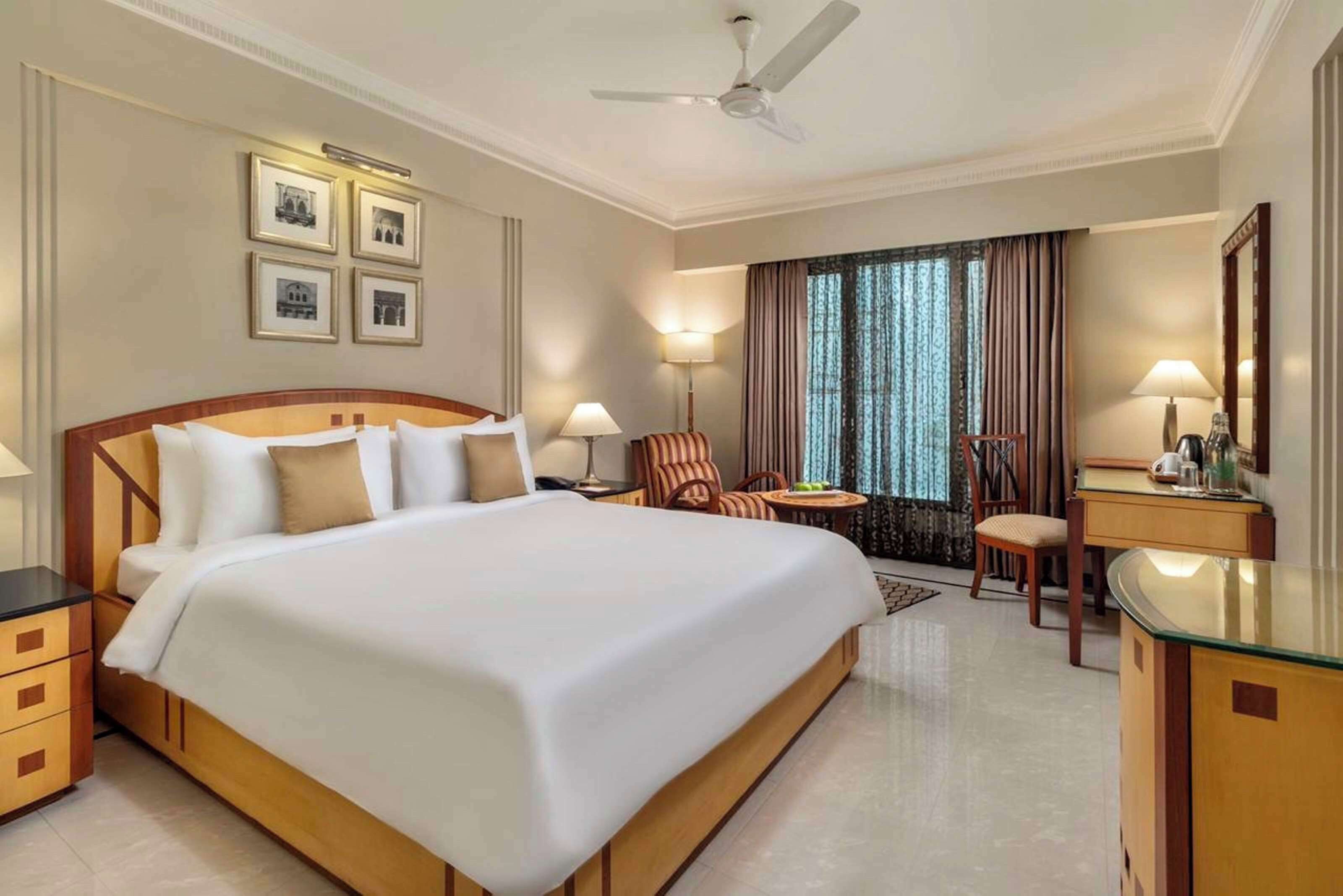 A hotel room with a large bed, a desk, a chair, and a small table. There are lamps, a ceiling fan, and framed pictures on the wall.
