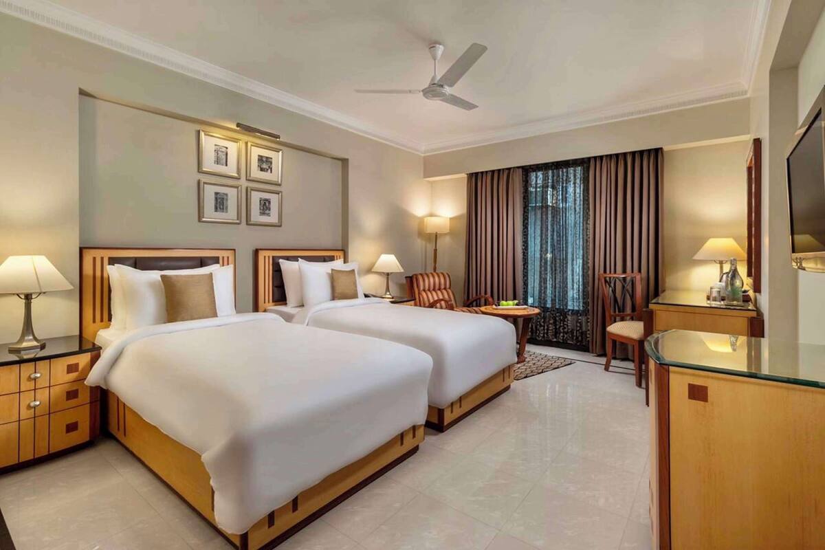 A hotel room with two beds, a desk, a chair, a TV, and a ceiling fan.
