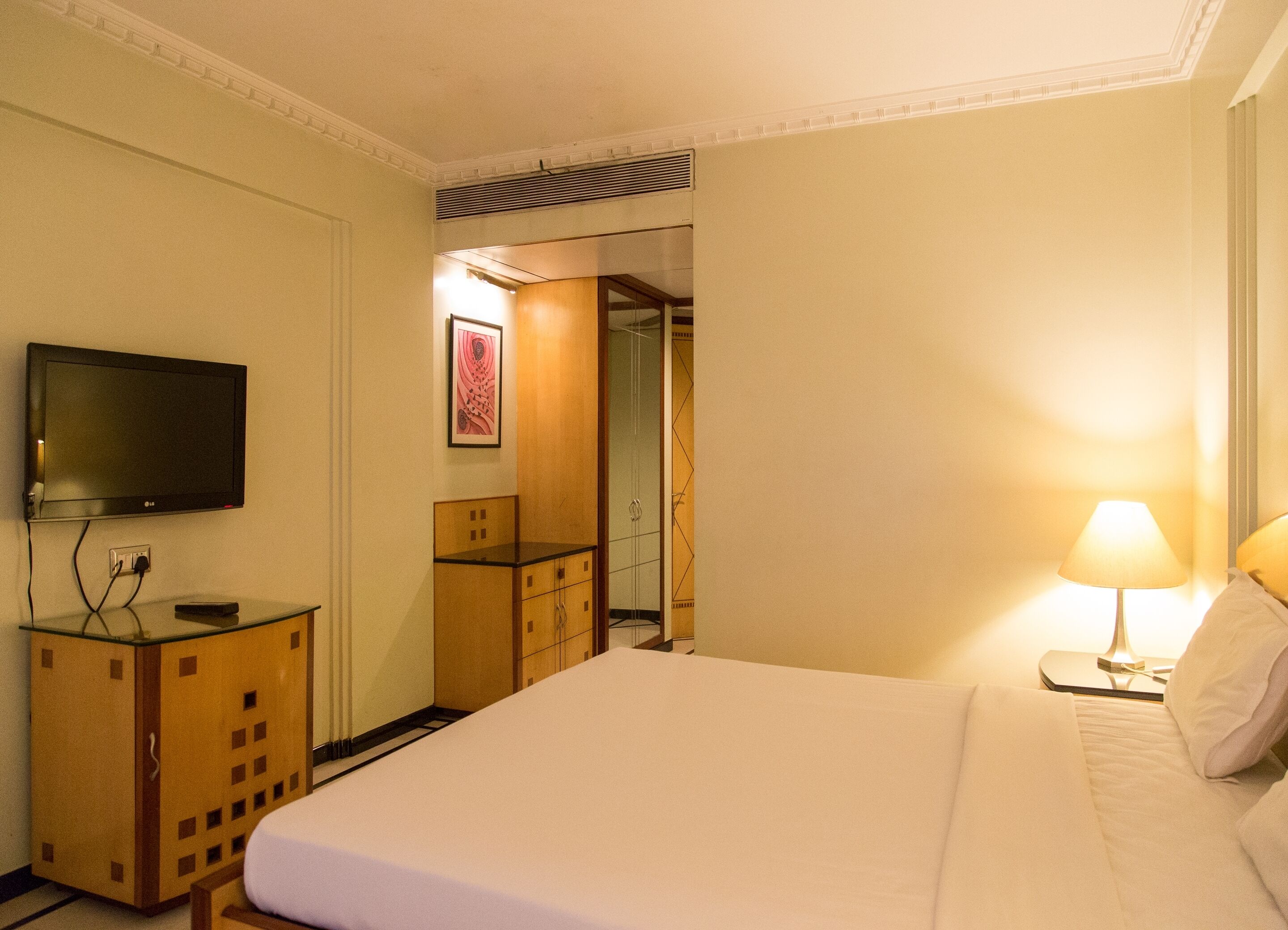A hotel room with a bed, a TV mounted on the wall, a desk with a lamp, and a door leading to another room.