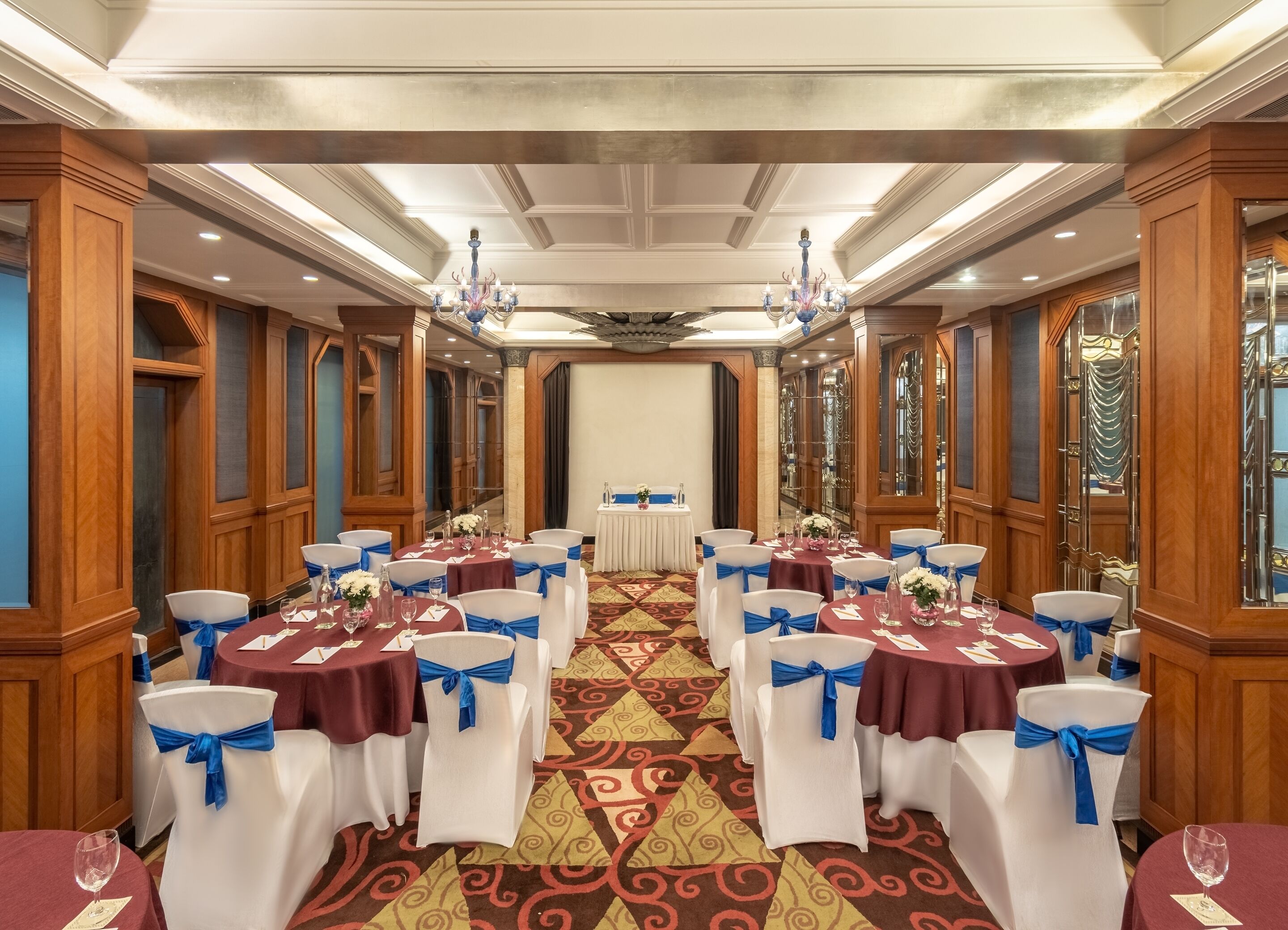 A banquet hall with round tables, white chairs with blue sashes, and a patterned carpet.