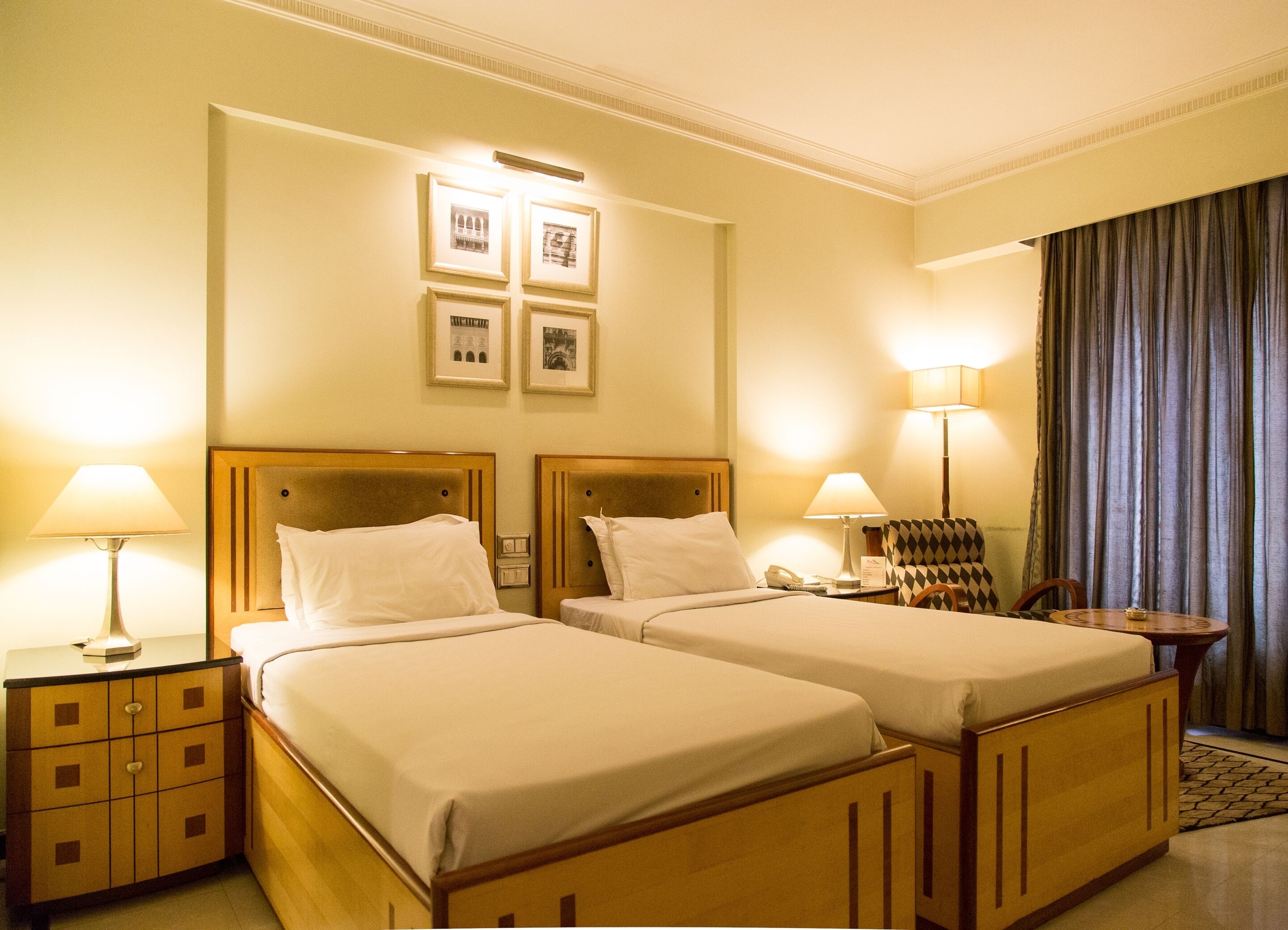 Une chambre d’hôtel avec deux lits simples, une table de chevet avec une lampe, une chaise et une petite table avec une lampe.