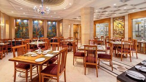 A spacious dining area with multiple tables and chairs, a chandelier, and artwork on the walls. - Sarovar Portico Kalupur Ahmedabad (Ahmedabad)