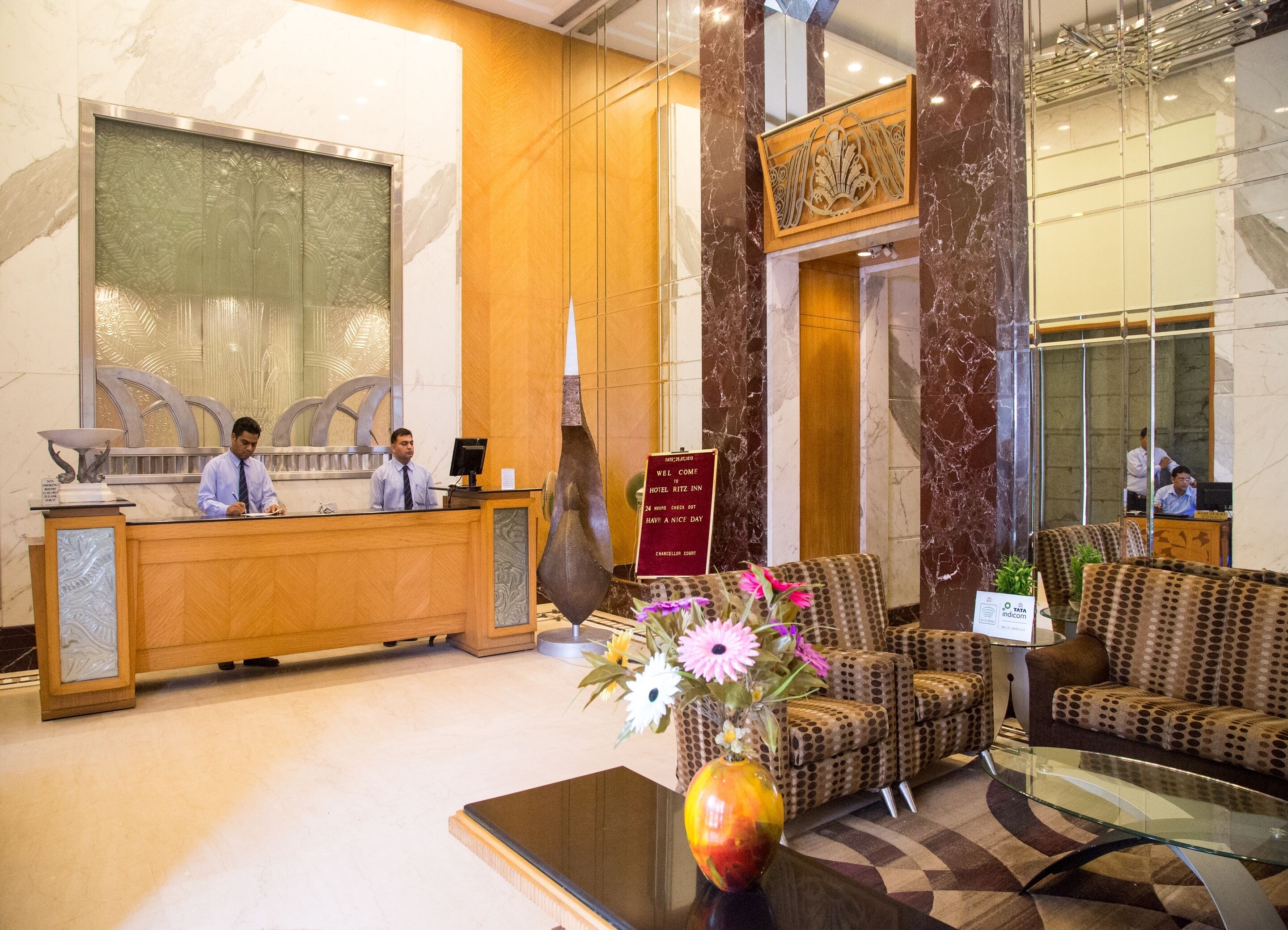 A hotel reception area with a wooden reception desk, a large decorative vase with flowers, and a seating area with patterned sofas.