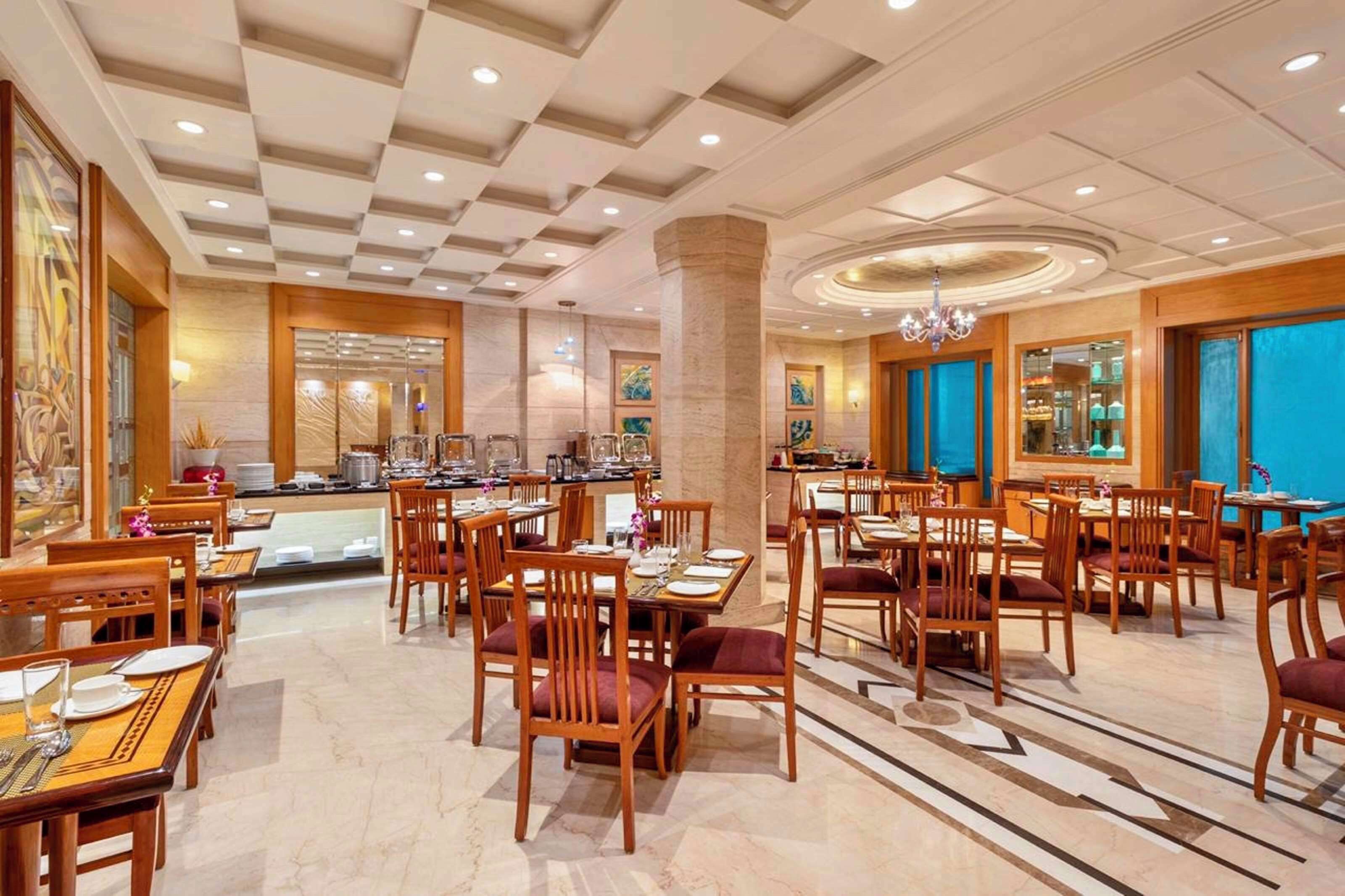 A hotel dining area with multiple tables and chairs, a central pillar, and a chandelier.