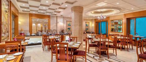 A hotel dining area with multiple tables and chairs, a central pillar, and a chandelier.
